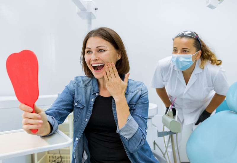 A person smiling into a mirror with a dental professional watching